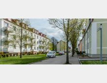 Straßenszene in einem Wohngebiet mit mehrstöckigen, hell gestrichenen Wohnhäusern auf beiden Seiten, teilweise mit Balkonen. Die Straße ist von Bäumen gesäumt, daneben befinden sich Rasenflächen, und parkende Autos stehen entlang der Fahrbahn.