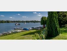 Grüne Uferlandschaft mit Rasenfläche und Bäumen, im Vordergrund kleine Bootsstege mit festgemachten Booten und im Hintergrund ein ruhiger Fluss mit mehreren Wasserfahrzeugen. Auf der anderen Seite des Flusses sind Häuser und eine von Bäumen gesäumte Küste zu sehen.