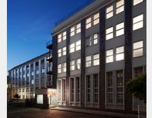 Moderne Bürogebäude mit mehreren Etagen, großen rechteckigen Fenstern und einer teilweise beleuchteten Fassade bei Dämmerung. Im Vordergrund befinden sich ein beleuchtetes Eingangsbereich mit einem Schild sowie ein Baum und ein gepflasterter Gehweg.