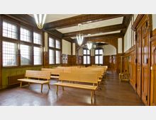 Ein Versammlungsraum mit dunklen Holzvertäfelungen, großen Fenstern mit Sprossenglas, Holzbänken in mehreren Reihen und bogenförmigen Deckenbalken. Hängelampen und eine Uhr an der Rückwand ergänzen die klassische Innenarchitektur des Raumes.