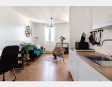 Wohnraum mit integrierter Küche in einem modernen, schlichten Stil. Links ein grünes Sofa mit Kissen, ein kleiner Tisch mit Pflanze und zwei Rattan-Stühle; rechts eine Küchenzeile mit weißen Schränken, Holzarplatte und Hängeschienen für Küchenutensilien.