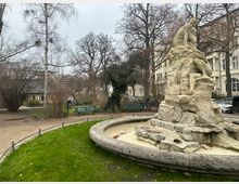 Kleiner Stadtpark mit einem trockenen Brunnen aus Stein, der eine Skulptur mit menschlichen Figuren zeigt. Im Hintergrund sind Bäume ohne Laub, eine Parkbank mit Graffiti und Gebäude im klassischen Stil zu sehen.
