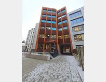 Ein modernes Bürogebäude mit einer rotbraunen Fassade und großen Glasfenstern. Der Haupteingang ist über Treppenstufen erreichbar, und rechts daneben führt eine überdachte Durchfahrt ins Gebäudeinnere. Der Boden ist teilverschneit.