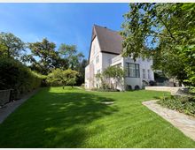 Großer Garten mit gepflegtem Rasen und vereinzelten Bäumen, umgeben von einer Hecke. Im Hintergrund ein zweistöckiges Haus mit hellrosa Fassade, hohen Fenstern und einer Terrasse aus Stein.