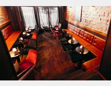 Innenraum einer Bar oder Lounge mit langen, braunen Leder-Sitzbänken entlang freiliegender Ziegelwände und mehreren kleinen runden Tischen mit Tischlampen. Dunkle Polsterstühle, Holzboden und große Fenster mit Vorhängen prägen den schmalen, länglichen Raum.