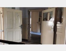 Eine verlassene Innenansicht mit mehreren beigen Holztüren, von denen einige beschädigt sind und kaputte Glasfenster haben. Der Raum wirkt alt und renovierungsbedürftig, mit abblätterndem Putz an den Wänden.