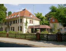 Zweistöckiges beige- und gelbfarbenes Gebäude mit roten Ziegeldächern und mehreren Fenstern mit braunen Fensterläden, umgeben von Bäumen und einem schmiedeeisernen Zaun. Im Vordergrund sind eine gepflasterte Straße und ein kleiner Garten zu sehen.