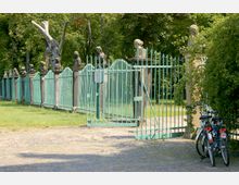 Eingang zu einem Park mit grünem Metallzaun und dekorativen Steinpostamenten, auf denen Büsten stehen, im Hintergrund Bäume und Grasflächen. Zwei Fahrräder lehnen rechts am Zaun.