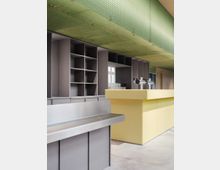 Moderner Innenraum mit einer gelben Theke und einer Rückwand mit grauen, offenen Regalfächern. Die Decke ist mit einem grünen, strukturierten Paneel verkleidet, und die Einrichtung umfasst Edelstahl-Oberflächen.