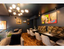 Gemütlicher Innenraum eines Cafés oder Restaurants mit dunkelblau gestrichenen Wänden, modernen Polsterstühlen und Tischen mit floral gemusterten Sitzbänken. An den Wänden hängen sowohl klassische Porträtgemälde als auch farbenfrohe, abstrakte Kunstwerke, und eine auffällige Deckenlampe mit kugelförmigen Leuchten erhellt den Raum.