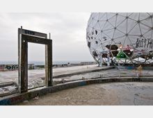 Verlassene Dachterrasse mit einer graffiti-bedeckten, halbkugelförmigen Radarkuppel und einer freistehenden, ebenfalls mit Graffiti bemalten Türrahmenstruktur. Der Boden ist teilweise beschädigt, und im Hintergrund ist ein bewölkter Himmel und eine weite Aussicht zu erkennen.