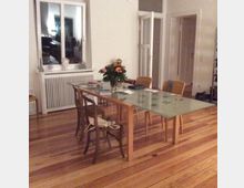 Ein Wohnzimmer mit Holzboden und einem Esstisch aus Glas, umgeben von mehreren Stühlen. Im Hintergrund ein Fenster mit weißem Heizkörper darunter sowie Türen, die zu anderen Räumen führen. Auf dem Tisch steht eine Blumenvase mit bunten Blumen.