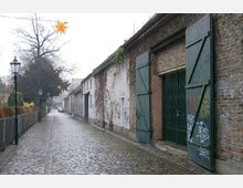 Kopfsteinpflasterstraße in einer ruhigen, städtischen Umgebung, gesäumt von alten Backsteinmauern, teilweise mit Graffiti und bewachsenen Fassaden. Im Hintergrund sind Bäume und eine gelbe, sternförmige Weihnachtsdekoration zu sehen.