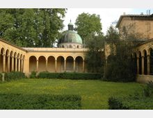Ein rechteckiger Klostergarten mit einer Wiese, blühenden gelben Blumen und gepflegten Hecken, umgeben von gelblichen Arkadengängen mit Rundbögen. Im Hintergrund ist eine große Kuppel mit grünlicher Patina zu sehen, umgeben von hohen Bäumen.