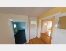 Flur eines Wohnhauses mit Holzboden, weißen Türen und einer gelb gestrichenen Wand. Zwei angrenzende Räume sind sichtbar, einer mit blau gestrichenen Wänden und dunklem Teppichboden, der andere mit weißen Wänden und großen Fenstern.