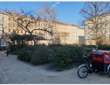 Ein kleiner innerstädtischer Park mit Kieswegen, kahlen Bäumen und Sträuchern. Im Hintergrund stehen helle Altbauwohngebäude, daneben ist ein Lastenfahrrad mit roter Abdeckung geparkt.