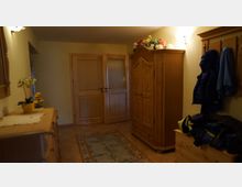 Flur in einem Wohnhaus mit gelb gestrichenen Wänden und hellen Holzmöbeln, darunter ein Schrank, eine Garderobe mit Jacken und ein kleiner Schubladenschrank mit einer gelben Topfpflanze. Der Boden ist mit beigefarbenen Fliesen und einem kleinen Teppich dekoriert.