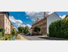 Straßenszene in einem Wohngebiet mit mehrstöckigen Backsteinhäusern auf der rechten Straßenseite, darunter ein Gebäude mit Satteldach und verzierten Fensterelementen. Links stehen kleinere, hell verputzte Häuser, ein Gehweg und eine grüne Hecke; der Himmel ist teils bewölkt.