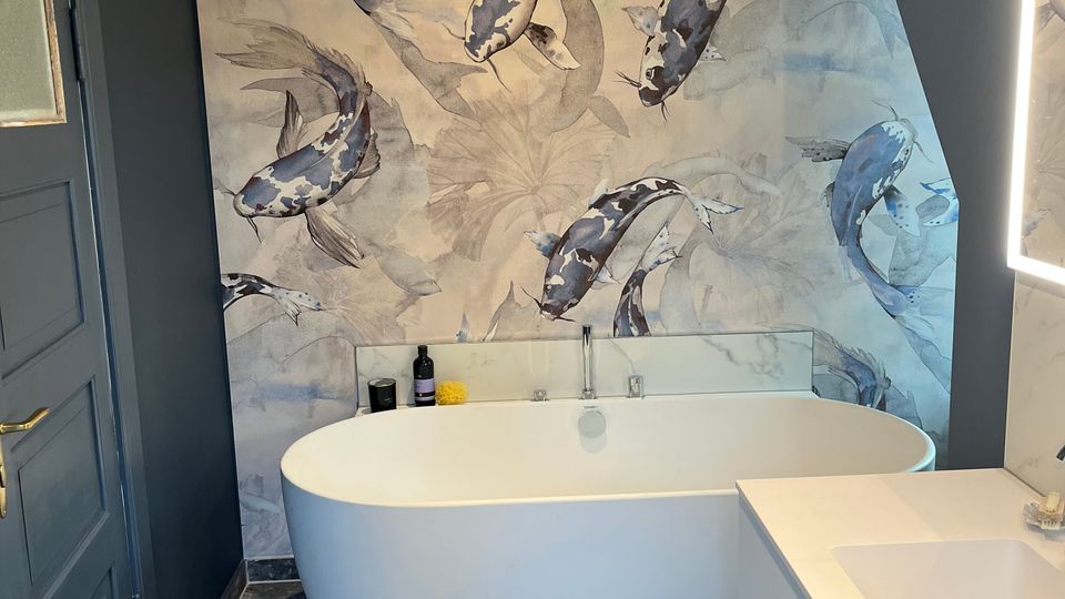 Modernes Badezimmer mit freistehender weißer Badewanne vor einer großen Wandfläche mit Koi-Fisch-Motivtapete. Dunkle Wände, graue Steinfliesen am Boden und ein weißer Waschtisch neben einem beleuchteten Spiegelrahmen prägen den Raum.