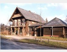 Ein zweistöckiges Blockhaus aus Holz mit Veranda und traditionellen Schnitzereien, daneben ein kleineres Nebengebäude mit Schrägdach. Das Haus liegt in einer ländlichen Umgebung mit einigen Bäumen und einem Gehweg im Vordergrund.