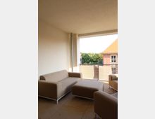 Ein überdachter Balkon mit beigen Polstermöbeln, bestehend aus einem Sofa, Sesseln und einem Hocker. Im Hintergrund befindet sich ein Geländer mit Blick auf ein Haus mit rotem Dach und grünen Bäumen.