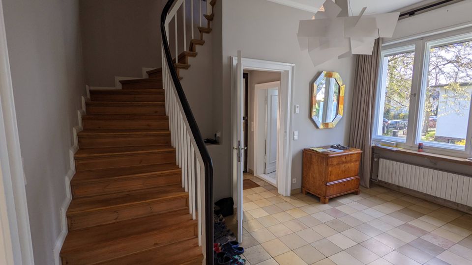 Heller Eingangsbereich eines Wohnhauses mit geschwungener Holztreppe und schwarzem Handlauf, beigen Fliesen sowie weiß-grauen Wänden. Rechts befinden sich große Fenster mit Vorhängen, ein Heizkörper, eine Holzkommode und ein achteckiger Wandspiegel; in der Mitte führt eine Tür in den Flur.