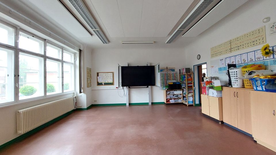 Klassenzimmer mit großen Fenstern an der linken Wand, die viel Tageslicht hereinlassen. Im Raum befinden sich eine digitale Tafel vorne, Regale voller Schulmaterialien an der rechten Wand und eine lange Heizkörperleiste unter den Fenstern.