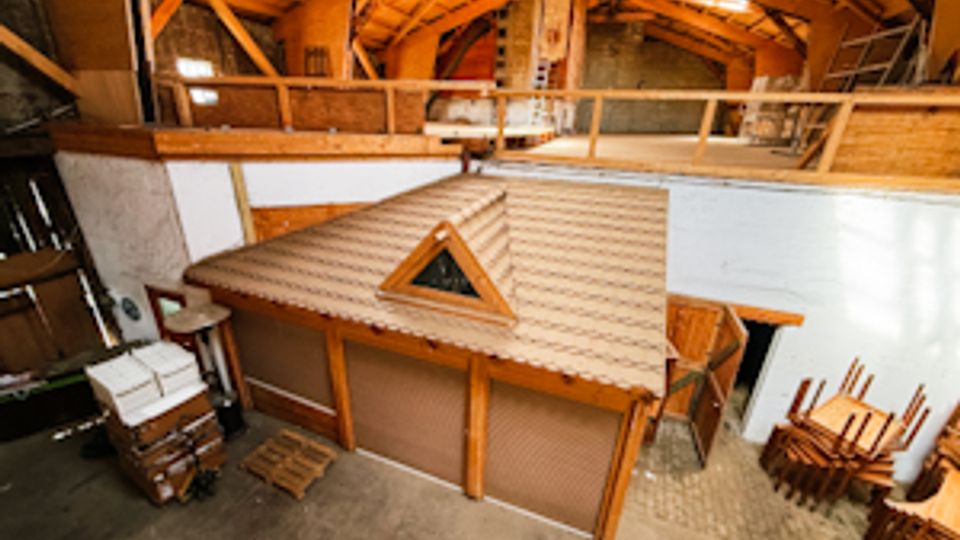 Innenansicht einer großen Scheune oder Lagerhalle mit hohem Holzdachstuhl und einer umlaufenden Galerie im Obergeschoss. Unten steht ein kleiner Holzbau mit Satteldach und Dreiecksfenster, umgeben von Betonboden, gelagerten Möbeln und Arbeitsmaterialien.