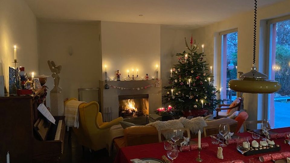 Gemütliches Wohnzimmer mit Kaminfeuer, dekoriert mit Kerzen und einem geschmückten Weihnachtsbaum neben großen Fenstern zur Terrasse. Im Vordergrund steht ein gedeckter Esstisch mit roter Tischdecke, Gläsern und Kerzen; links sind ein Klavier und Sitzmöbel zu sehen.