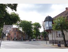 Straßenszene in einer Kleinstadt mit einer Wegweiser-Tafel, die zu einer Musikakademie, einem Schloss und dem Rathaus zeigt. Im Hintergrund sind historische Gebäude, darunter ein Haus mit Erker und einem grauen Schieferdach, sowie Bäume und eine Ampelkreuzung zu sehen.