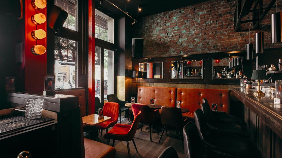 Innenraum einer Bar mit hohen Fensterfronten, sichtbarer Backsteinwand und dunkler Decke mit Spotbeleuchtung. Entlang der Bar und einer gepolsterten Sitzbank stehen kleine Tische mit gemischten Stühlen; Spiegel und Hängelampen rahmen den Tresenbereich.