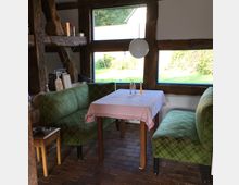 Gemütliche Sitzecke in einem rustikalen Raum mit Holzbalken und Ziegelboden. Zwei grüne Sofas mit kariertem Muster stehen um einen kleinen Tisch mit rosa Tischdecke, vor großen Fenstern mit Blick auf einen Garten.