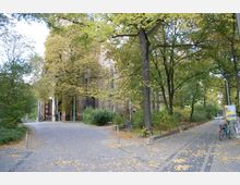 Gepflasterter Weg in einem parkähnlichen Bereich, gesäumt von hohen Bäumen mit herbstlichem Laub. Im Hintergrund ist ein historisches Gebäude mit Backsteinfassade und Torbogen sichtbar, während im Vordergrund Fahrräder an Laternen und Geländern lehnen.