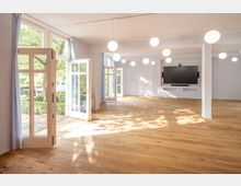 Großer, heller Raum mit Holzboden, großen Fenstern und Glastüren, die ins Grüne führen. An der weißen Wand ist ein großer moderner Bildschirm angebracht, und mehrere runde Hängelampen beleuchten den Raum.