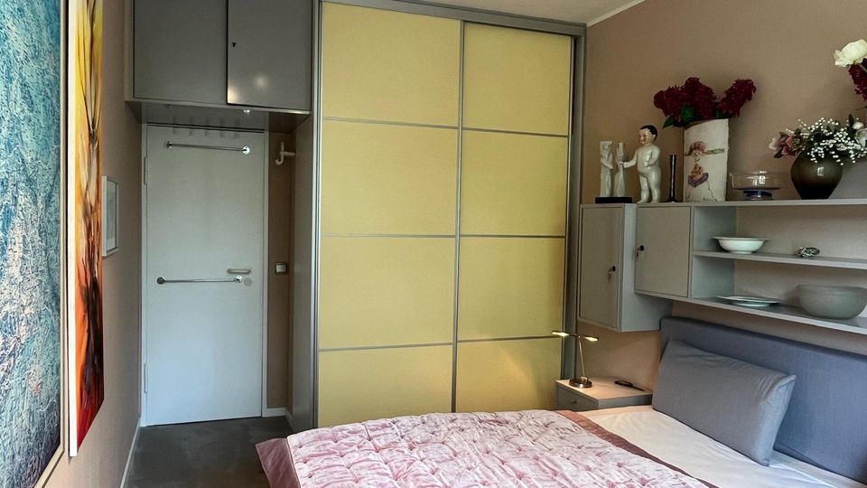 Schmales, modern eingerichtetes Schlafzimmer mit Doppelbett und rosa Tagesdecke, seitlichen Regalen und Wanddekorationen. Am Ende des Raums stehen eine weiße Tür sowie ein großer Schiebetür-Kleiderschrank mit hellgelben Paneelen und grauem Rahmen.