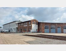 Ziegelgebäude eines historischen Lokschuppens mit großen blauen Toren und aufgesetzten Schildern wie „Lokdienstleitung“ und „Abteilung Tb“. Daneben ein weißes, längliches Gebäude; im Vordergrund verlaufen Gleise und ein asphaltierter Gehweg.