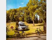 Wohnmobil auf einer Wiese unter hohen Bäumen, mit einer ausgeklappten Markise und Campingstühlen. Im Vordergrund stehen mehrere Fahrräder, und im Hintergrund befindet sich eine baumbestandene Parklandschaft bei sonnigem Wetter.