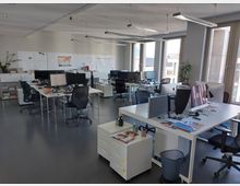 Modernes Großraumbüro mit mehreren weißen Schreibtischen, versehen mit Monitoren, Bürostühlen und Arbeitsmaterialien. Helle Fensterfront und minimalistische Einrichtung mit einigen Pflanzen sorgen für eine offene und lichtdurchflutete Atmosphäre.