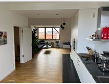 Moderne Wohnung mit offener Küche und Wohnbereich, in hellem Design gehalten. Zu sehen sind eine Einbauküche mit dunkler Arbeitsplatte rechts, ein heller Holzfußboden, ein weißer Wandanstrich und ein Wohnzimmer mit großen Fenstern, die Tageslicht hineinlassen.