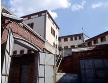 Backstein-Gebäudekomplex mit weiß verputzten höheren Strukturen, Metalltüren und einem Außenbereich mit Treppe; Industrie- oder Lagerhaus-Architektur, wahrscheinlich in einem älteren urbanen Umfeld.