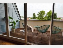 Blick durch eine Glastür auf einen Balkon mit Holzboden, der mit zwei grünen Stühlen, einem Glastisch und einer Pflanze in einem Topf dekoriert ist; im Hintergrund sind Bäume und umliegende Gebäude zu sehen.