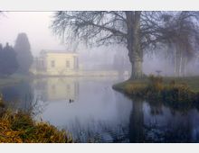 Neblige Parklandschaft mit einem kleinen See, einem großen kahlen Baum im Vordergrund und einem klassizistischen Gebäude mit Säulen im Hintergrund. Am Ufer ist Herbstlaub zu sehen, während sich das Wasser ruhig spiegelt.