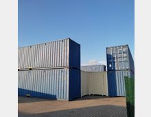 Stapel von Frachtcontainern in Blau und Beige auf einem gepflasterten Lagerplatz bei klarem Himmel. Die Container sind unterschiedlich hoch gestapelt, und ein Teilbereich eines grünen Zauns ist am rechten Bildrand sichtbar.