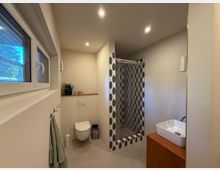 Modernes Badezimmer mit neutralen Beigetönen; ausgestattet mit einer bodenebenen Dusche mit gemusterten Fliesen, einem wandmontierten WC, einem weißen Waschbecken auf einem Holzunterschrank und einem schmalen Fenster auf der linken Seite.