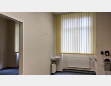 Leerer Raum mit beigem Anstrich, einer hohen Fensterfront mit gelben Lamellenjalousien, einem kleinen Waschbecken an der Wand und einer weißen Heizung darunter. Auf der rechten Seite befinden sich Kabel und Wandsteckdosen.