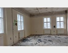 Ein leerer Raum mit cremefarbenen Wänden und vier großen Fenstern mit weißen Rahmen. Der Boden zeigt Abnutzungsspuren, und an den Wänden sind Heizkörper sowie einige offene Kabel und Wanddurchführungen sichtbar.