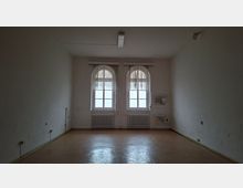 Leerer, schlicht gehaltener Innenraum mit weißen Wänden und braunem Boden, beleuchtet durch zwei große Rundbogenfenster an der Stirnwand. Unter den Fenstern stehen Heizkörper; rechts ist ein kleines Wandwaschbecken mit Ablage und sichtbaren Anschlüssen montiert.