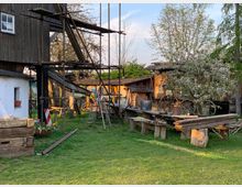 Hinterhof mit Rasenfläche, alten Holzbauten und einem blühenden Baum. Links steht ein teilweise eingerüstetes, holzverkleidetes Gebäude, daneben liegen Bretter, Rohre und Werkzeuge auf einfachen Holztischen und Brücken. Im Hintergrund stehen verwitterte Holzwagen und eine Scheune.