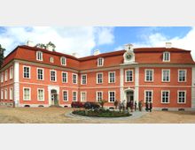 Rotes barockes Herrenhaus mit weißen Fensterrahmen und einem Uhrengiebel, umgeben von einem gepflasterten Vorplatz und einem kleinen Blumenbeet; vor dem Gebäude stehen Menschen und parkende Autos.