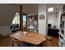 Heller Wohnbereich mit offener Gestaltung, Holzboden und modernen Möbeln. Im Vordergrund ein Esstisch mit Stühlen und einer Blumenvase, dahinter ein Bereich mit Backsteinwand, Regalen und großen Fenstern, die auf eine Terrasse führen.